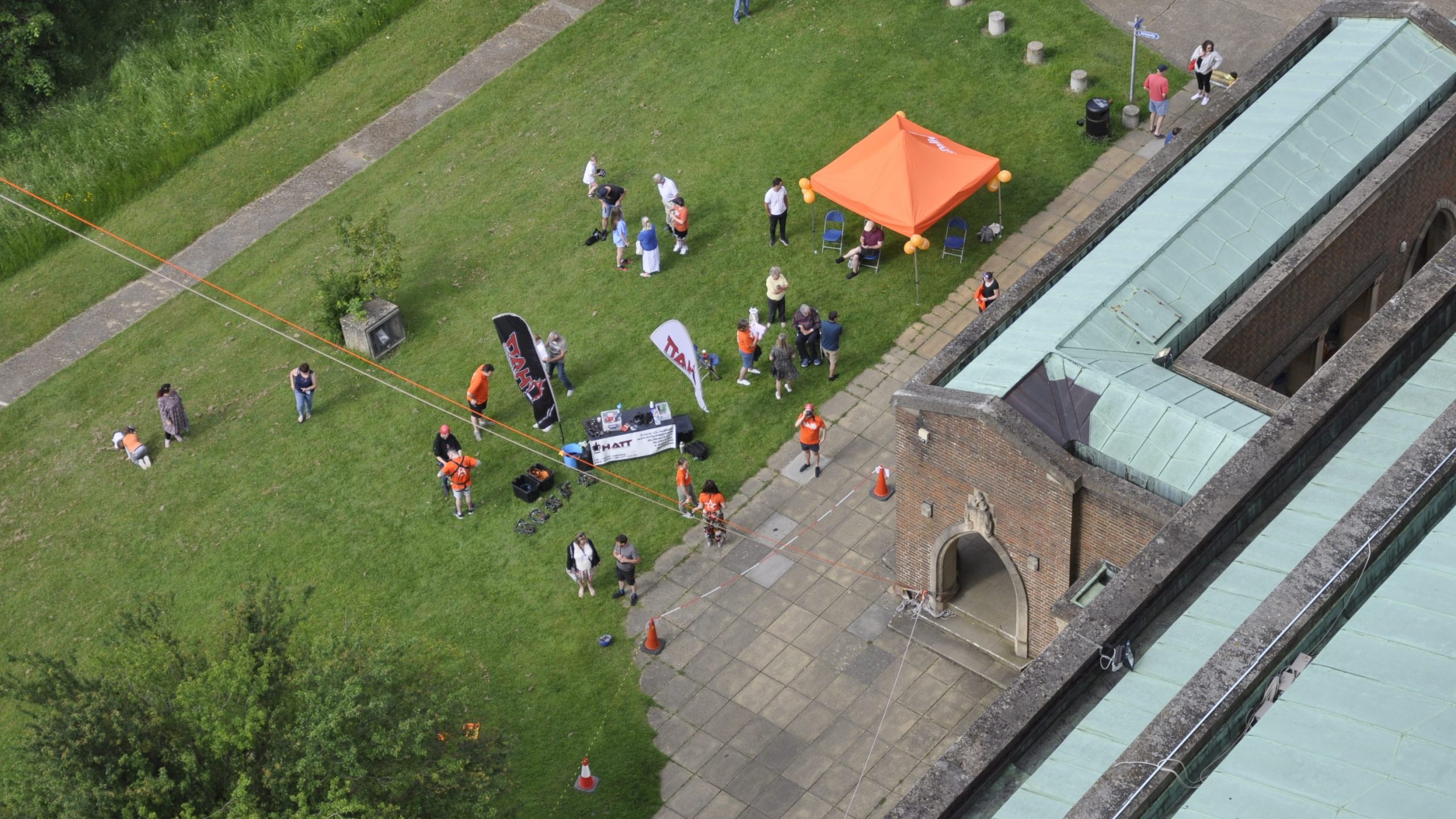 View from the top of an abseil, down to grassy ground. There is an orange gazebo, a sign up desk and a crowd of approximately 25 people gathered.