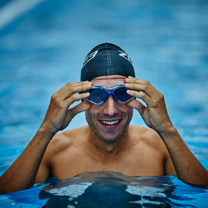 A man wearing a zone 3 blue framed goggles in a pool also wearing a black zone 3 swimming cap as well.